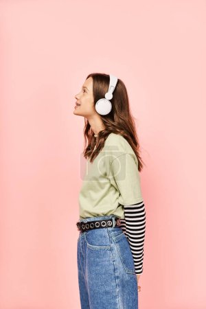 A stylish teenage girl in vibrant attire stands against a pink wall, wearing headphones and posing actively.