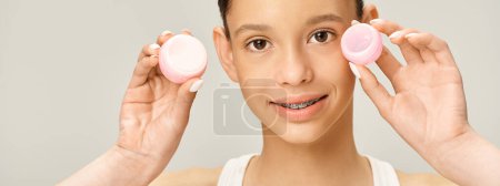 Foto de Una elegante adolescente sosteniendo crema frente a su cara, posando activamente con un atuendo vibrante. - Imagen libre de derechos