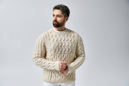A captivating man with a beard strikes a pose in a white sweater against a grey background in a studio setting.