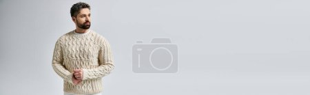 A mysterious man with a beard striking a pose in a white sweater against a grey studio background.