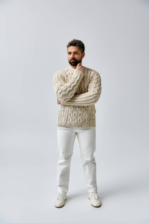 Foto de Un hombre barbudo exuda sofisticación en un suéter blanco y pantalones contra un fondo gris del estudio. - Imagen libre de derechos