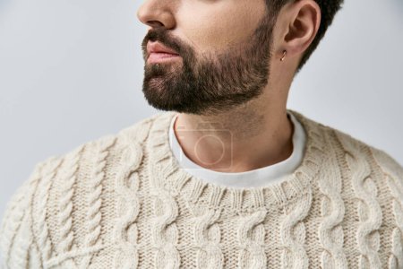 Ein stilvoller Mann mit Bart posiert in einem gemütlichen weißen Pullover vor schickem grauen Hintergrund in einem Studio.
