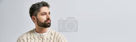 Un homme charismatique avec une barbe frappe une pose dans un pull blanc confortable sur un fond de studio gris.
