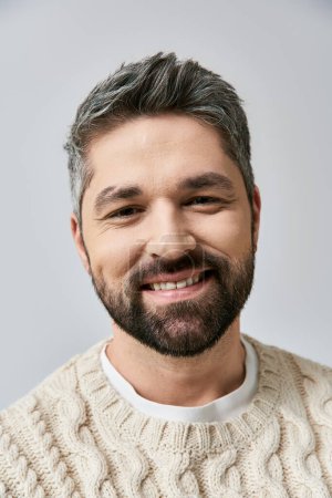 Foto de Un hombre sereno, de pelo gris y barba, vestido con un suéter blanco, sonríe calurosamente contra un fondo gris del estudio. - Imagen libre de derechos