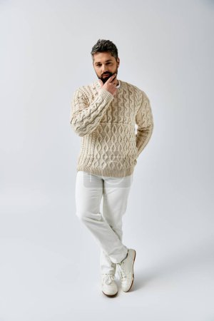 Foto de Un hombre barbudo posa elegantemente en un suéter blanco sobre un fondo gris del estudio. - Imagen libre de derechos