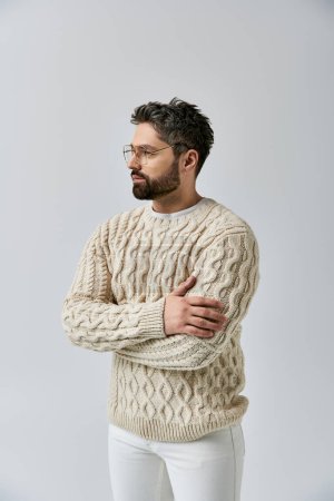 A bearded man strikes a captivating pose in a white sweater and pants against a grey studio backdrop.