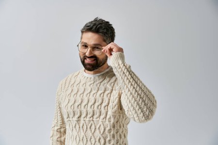 Foto de Un hombre barbudo exuda encanto en un suéter blanco y gafas sobre un fondo gris estudio. - Imagen libre de derechos
