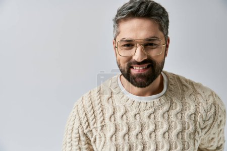 A bearded man exudes allure in a white sweater, complemented by glasses, on a grey backdrop in a studio setting.