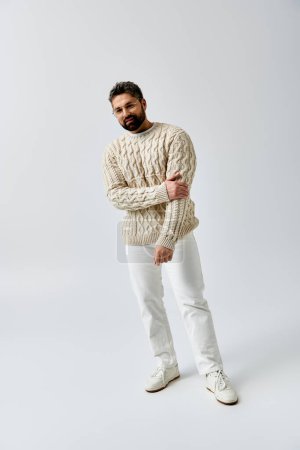 Foto de Un hombre carismático con barba posa en un suéter blanco de moda sobre un simple fondo gris de estudio. - Imagen libre de derechos
