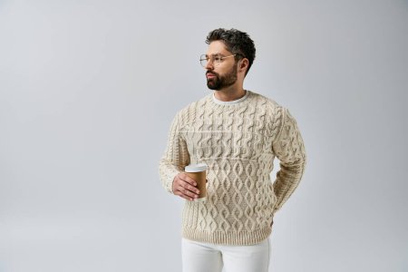 Foto de Un hombre barbudo emana encanto en un suéter blanco sobre un fondo gris, mostrando un aspecto sofisticado y pulido. - Imagen libre de derechos