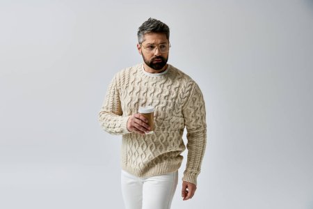 Foto de Un hombre cautivador con barba sosteniendo elegantemente una taza de café, exudando tranquilidad y relajación en un suéter blanco sobre un elegante fondo gris. - Imagen libre de derechos
