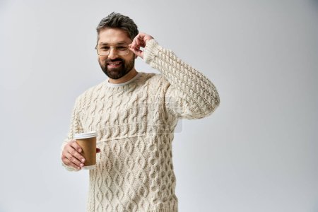 Un homme barbu dans un pull blanc tient délicatement une tasse de café fumante sur fond de studio gris.