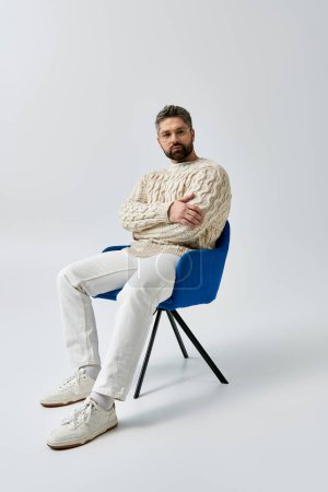 Foto de Un hombre barbudo en un suéter blanco se sienta con los brazos cruzados, exudando encanto y contemplación sobre un fondo gris del estudio. - Imagen libre de derechos