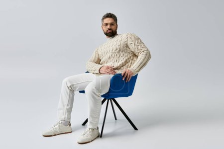Ein stilvoller Mann mit Bart sitzt auf einem blauen Stuhl in einem Studio mit grauem Hintergrund..