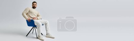 Photo for A bearded man in a white sweater sits gracefully in a chair, exuding an air of contemplation and sophistication. - Royalty Free Image
