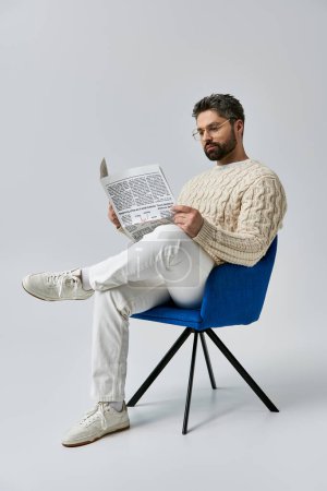 A bearded man in a white sweater sits in a chair, engrossed in reading a newspaper against a grey background.