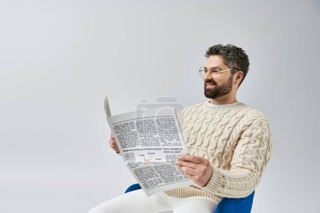 A stylish man with a beard sits on a chair, engrossed in reading a newspaper, against a grey background in a studio.