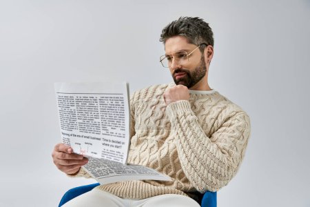 Un homme barbu dans un pull blanc est assis sur une chaise, absorbé par la lecture d'un papier sur un fond de studio gris.