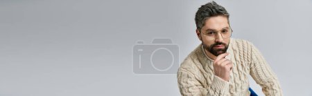 Photo for A bearded man in a white sweater sits on a chair, deep in thought with his hand resting on his chin, against a grey background. - Royalty Free Image