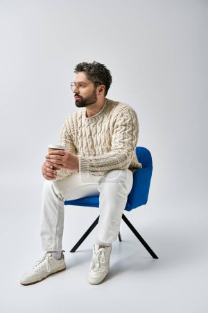 Téléchargez les photos : Un homme barbu dans un pull blanc s'assoit sur une chaise bleue, croisant ses bras dans une pose contemplative sur un fond gris. - en image libre de droit