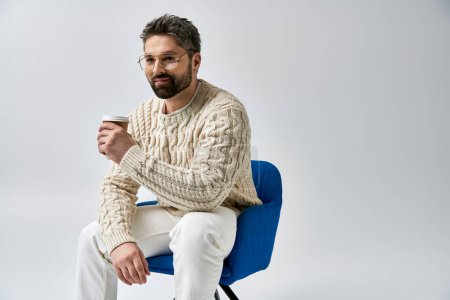 Photo for A man with a beard, dressed in a white sweater, sits on a chair holding a cup of coffee against a grey studio background. - Royalty Free Image