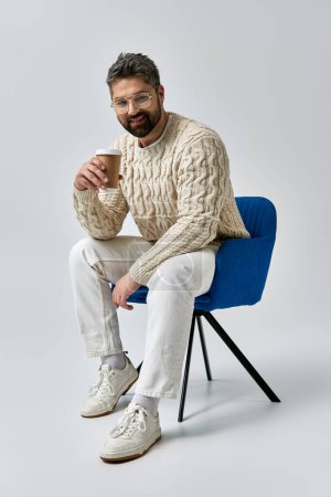 Photo for A stylish man with a beard sits on a chair in a white sweater, savoring a cup of coffee on a grey studio backdrop. - Royalty Free Image