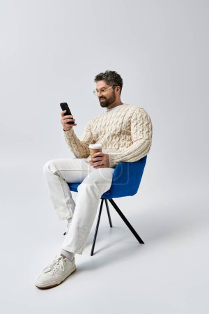 Foto de Un hombre barbudo en un suéter blanco se sienta pacíficamente en una silla, absorto en su teléfono celular. - Imagen libre de derechos