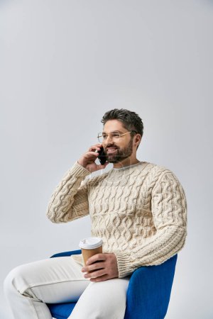 Foto de Un hombre elegante con barba se sienta en una silla, enfocado en su teléfono celular mientras está ocupado en una conversación telefónica. - Imagen libre de derechos