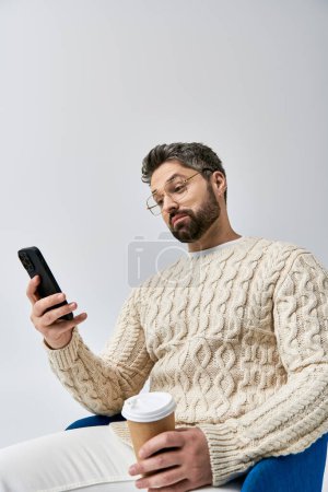 Foto de Un hombre barbudo en un suéter blanco se sienta en una silla, absorto en su teléfono celular. - Imagen libre de derechos