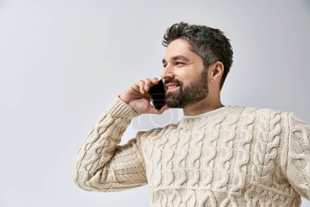 Téléchargez les photos : Un homme élégant avec une barbe parlant sur un téléphone portable tout en portant un pull blanc sur un fond gris dans un studio. - en image libre de droit