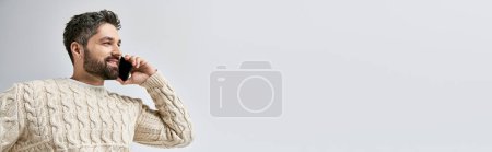 Photo for A bearded man in a white sweater is talking on a cell phone in a serene manner. - Royalty Free Image