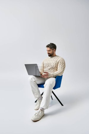 A bearded man in a white sweater sits on a chair, engrossed in his laptop on a grey background in a studio setting.