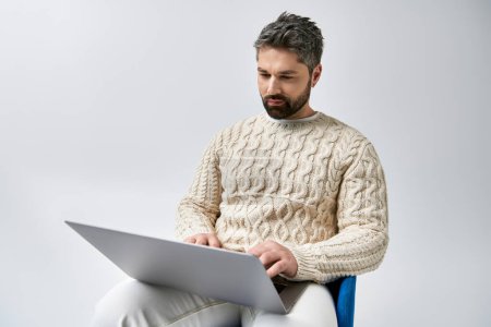 Foto de Un hombre cautivador con una barba absorta en el uso de un ordenador portátil mientras está sentado en una silla contra un fondo de estudio gris. - Imagen libre de derechos