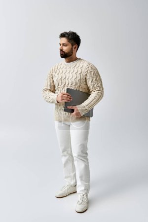 Foto de Un hombre barbudo con un suéter blanco sosteniendo un portátil en una pose cautivadora sobre un fondo gris. - Imagen libre de derechos