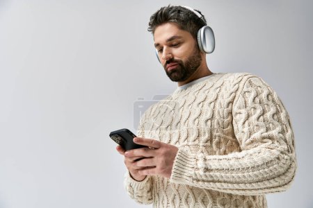 A bearded man wearing headphones gazes at his phone, lost in the music playing through his headset.