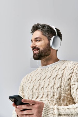 Ein bärtiger Mann im weißen Pullover trägt Kopfhörer, in seinen Handybildschirm vor grauem Hintergrund vertieft.