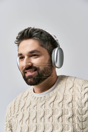Téléchargez les photos : Un homme captivant avec une barbe coiffée d'écouteurs et un pull blanc, plongé dans le rythme de la musique. - en image libre de droit