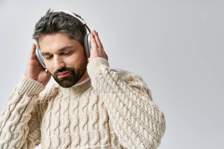 Foto de Un hombre elegante con barba lleva un suéter blanco, inmerso en la música a través de auriculares, sobre un fondo gris del estudio. - Imagen libre de derechos