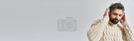 Téléchargez les photos : Un homme barbu dans un élégant pull blanc tient un téléphone portable à l'oreille sur un fond de studio gris. - en image libre de droit