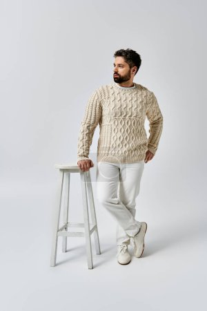 A stylish man with a beard stands confidently next to a stool, wearing a fashionable white sweater in a studio setting.