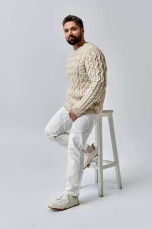 Un homme barbu respire le charme en s'asseyant sur un tabouret, élégamment vêtu d'un pull blanc sur fond de studio gris.