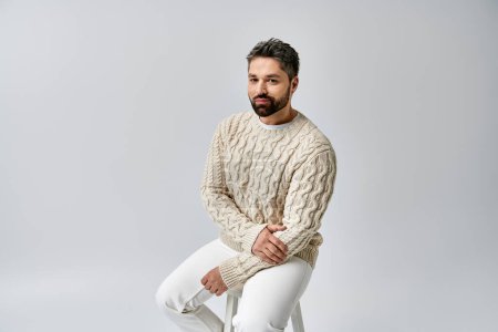 A charismatic man with a beard sits on a stool, exuding charm in a cozy white sweater against a grey studio backdrop.