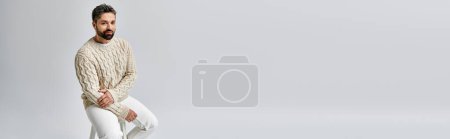 Téléchargez les photos : Un homme barbu au regard séduisant pose dans un pull blanc et un pantalon sur fond de studio gris. - en image libre de droit