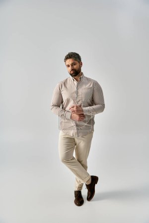 A fashionable man with a beard wearing a striped shirt and khaki pants poses elegantly on a grey background in a studio.