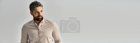 A stylish man with an alluring beard poses confidently in elegant attire against a sleek white backdrop.