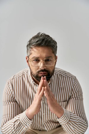 A stylish man with glasses and a beard sits gracefully, lost in thought on a grey background.