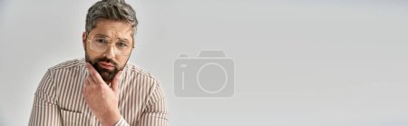 Téléchargez les photos : Un homme élégant avec une barbe et une tenue élégante semble surpris dans un cadre de studio sur un fond gris. - en image libre de droit