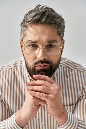 Ein stilvoller Mann mit Bart, Brille und gestreiftem Hemd posiert elegant vor grauem Studiohintergrund.