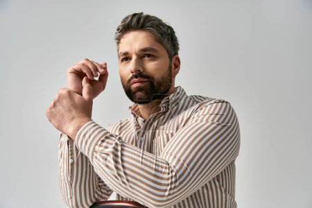 Foto de Un hombre elegante con barba posa con una camisa a rayas sobre un fondo gris de estudio. - Imagen libre de derechos