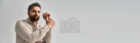 Téléchargez les photos : Un homme barbu dans une élégante chemise rayée tient ses mains ensemble, exsudant sophistication et style sur fond de studio gris. - en image libre de droit
